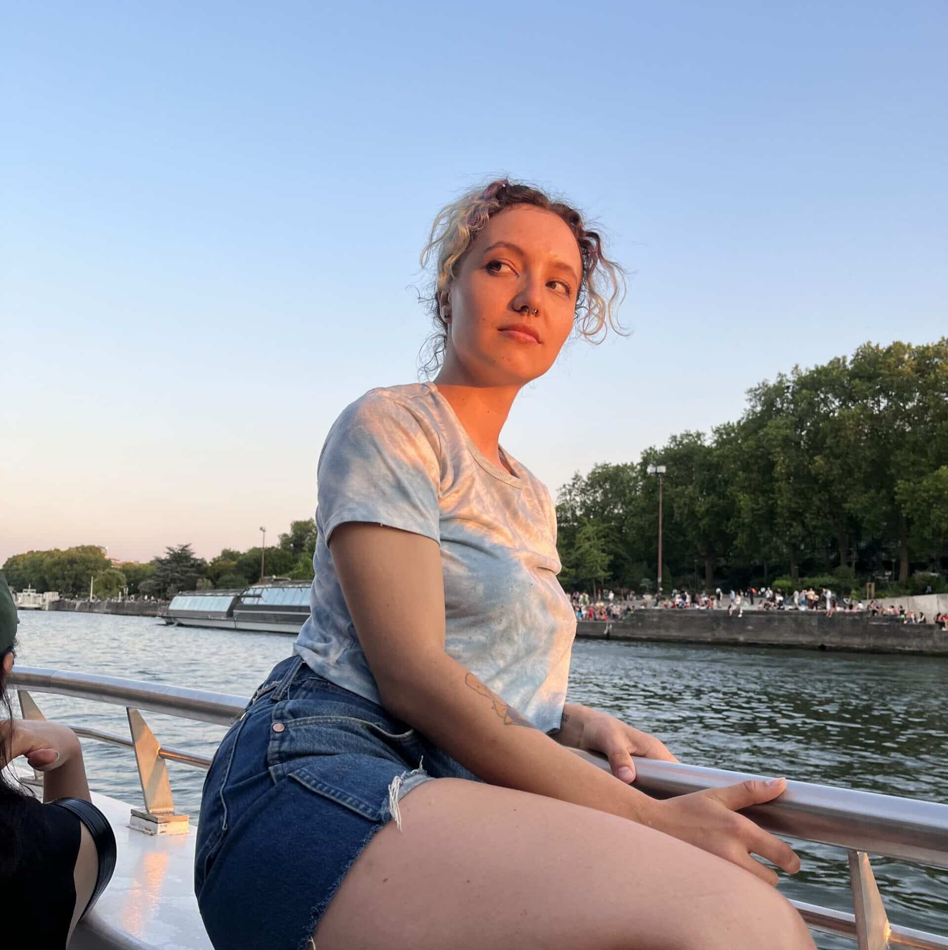 I am on a boat tour in Paris. I am wearing a blue tie-dye shirt and jean shorts. My hair is blue-gray.