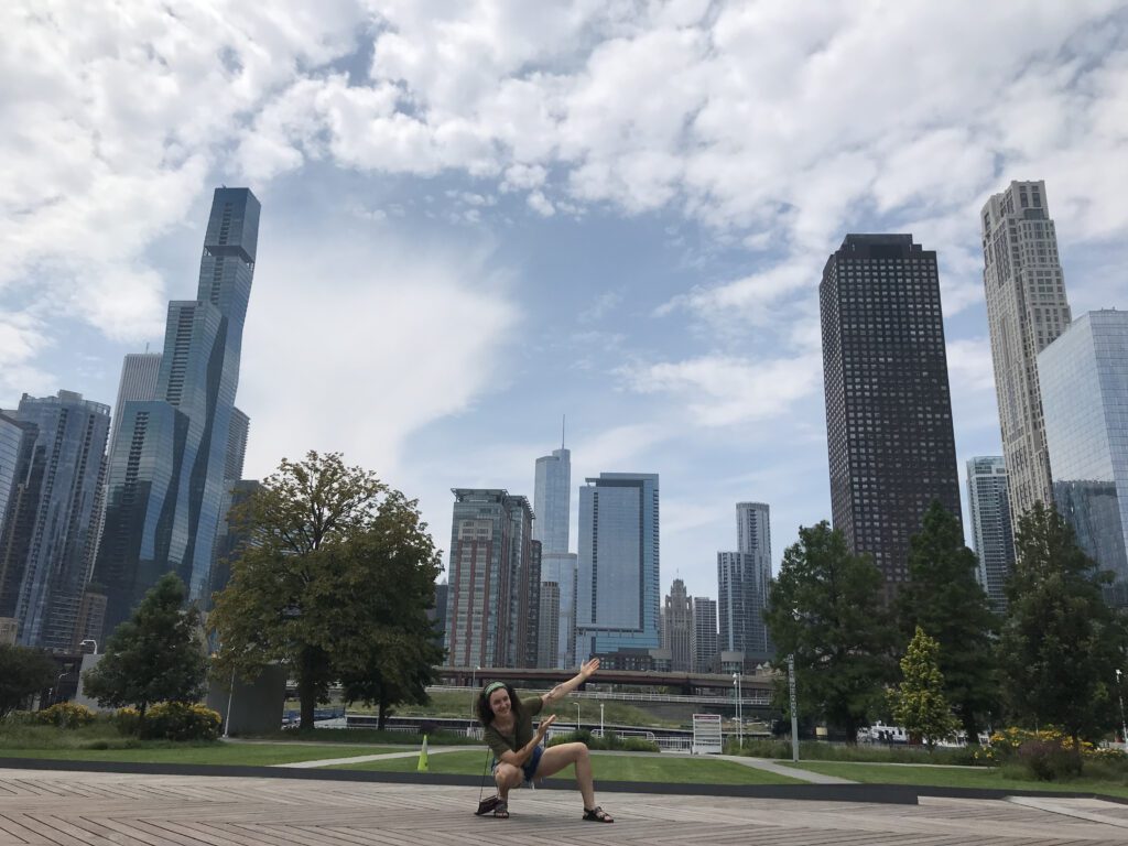 I am crouched in front of the Chicago cityscape. I am gesturing behind me. My hair is brown and I'm wearing a green headband.