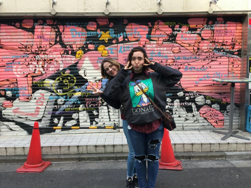 I am standing in front of a mural in Tokyo, Japan with a friend. I have brown hair that is in a beanie. I am making peace signs.