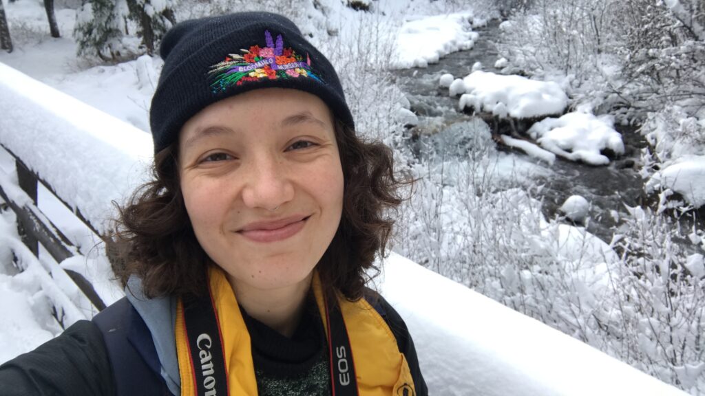 I am standing on a snow-covered bridge in Oregon. I am wearing a beanie and my hair is brown. I am smiling.