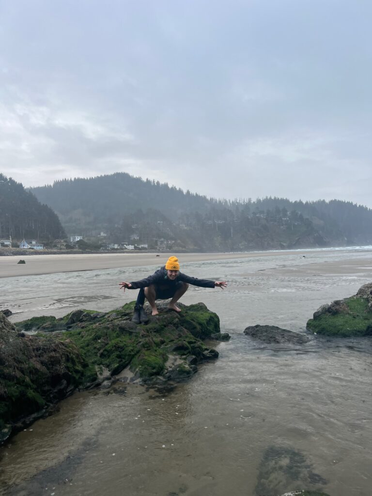 I am crouched on a rock at the coast of Oregon. I do not have shoes on. My hair is up in buns and I am wearing a beanie. My hair is blue-gray.