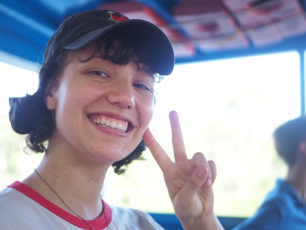 I am smiling and holding up a peace sign. My hair is brown, up in buns, and I'm wearing a hat.